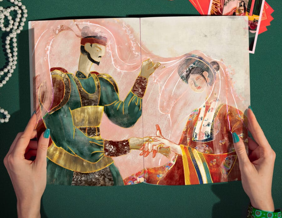 A person opening a vibrant book with thick, glossy paper. The book spread displays an illustration of two people dressed in vibrant attire