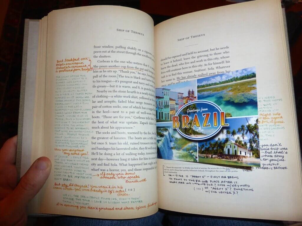 Person opening a book with wide margins, filled with handwritten notes in various handwriting and pen colors, and a postcard nestled between the pages
